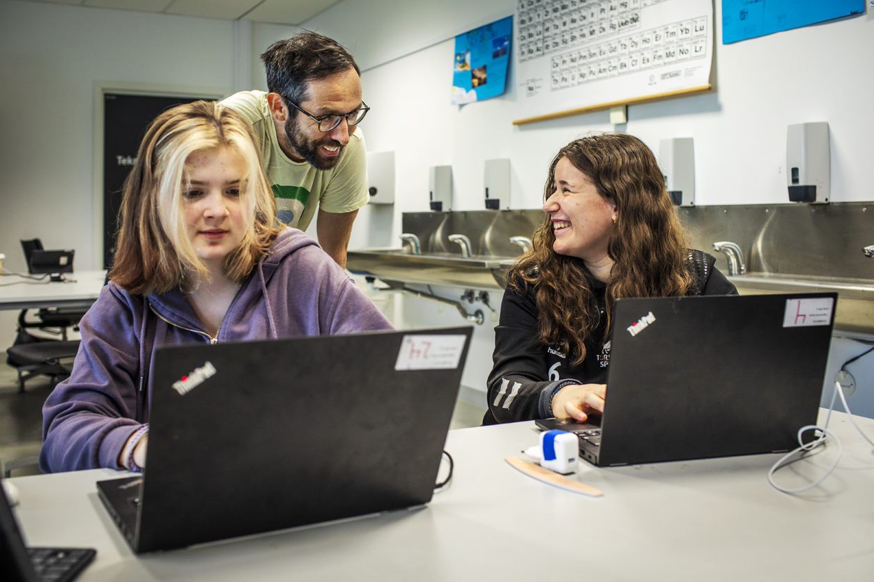 To elever som sitter ved hver sin PC i klasseromet får veiledning av en lærer